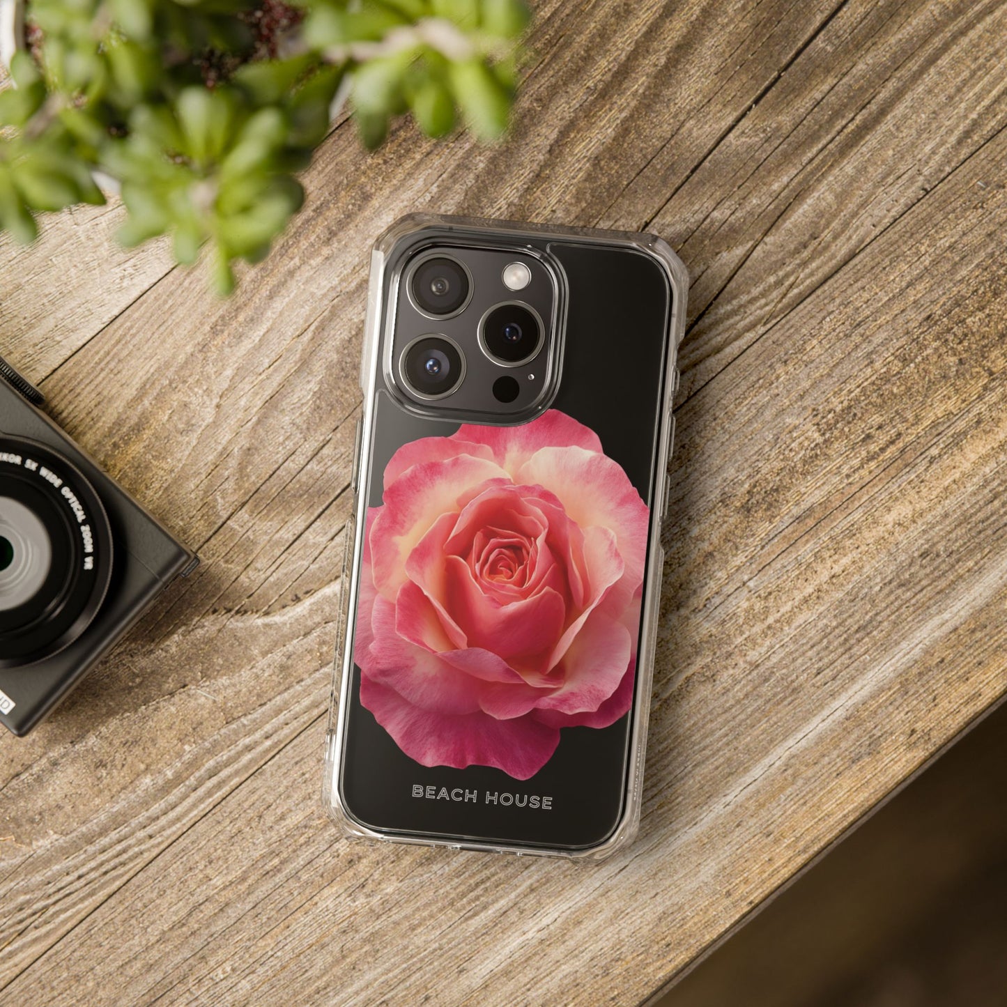 Phone case with pink rose design on a wooden surface next to a camera and plant.