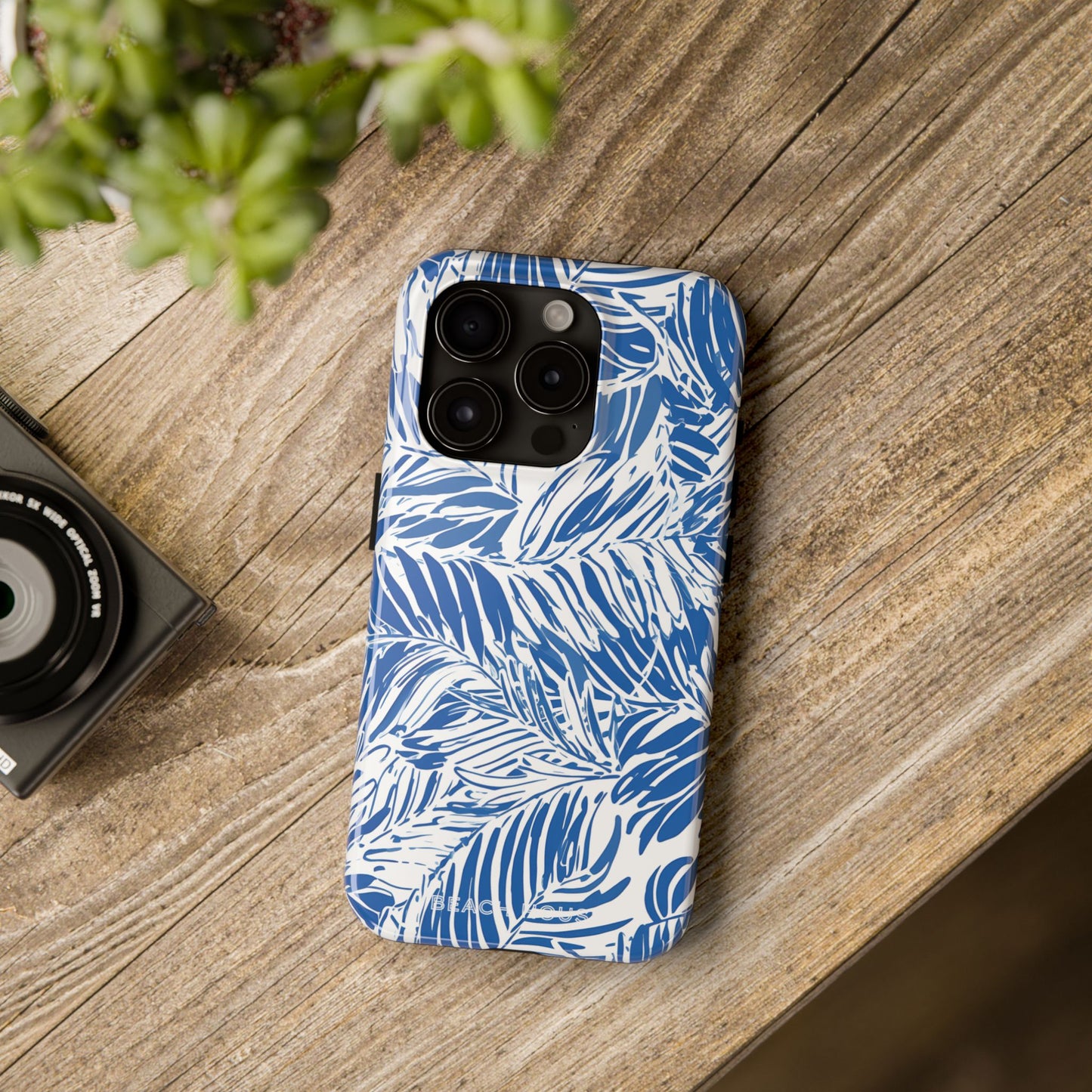 Blue and white palm leaf patterned Hampton Tough Case for iPhone on a wooden surface with a camera and plant in the background.
