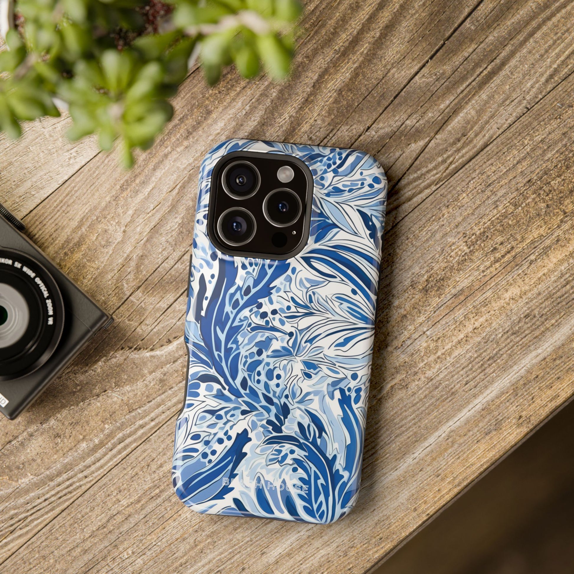 Blue and white floral patterned Nags Head MagSafe Tough Case for iPhone on a wooden surface with a camera and plant in the background.