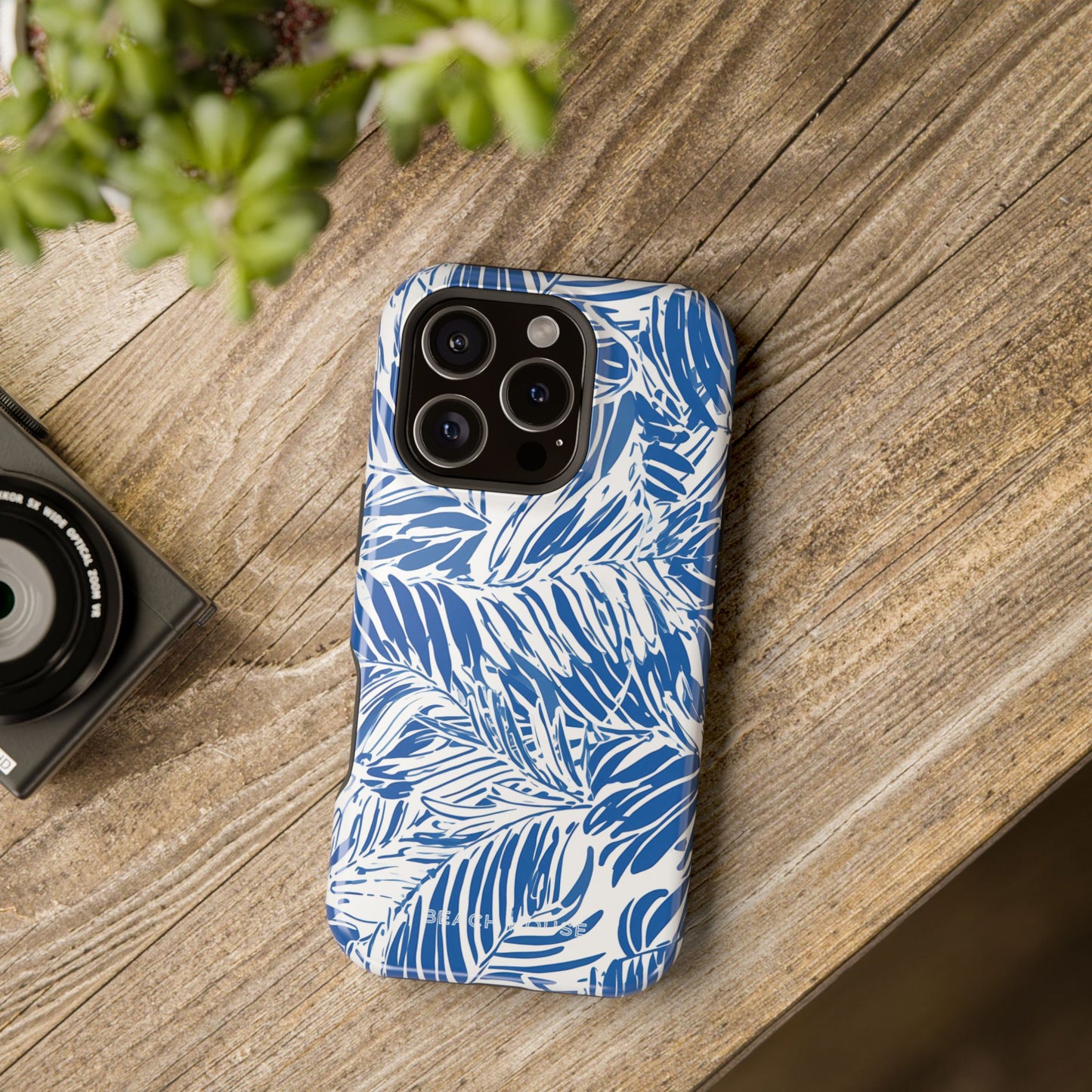 Blue and white Hampton MagSafe Tough Case for iPhone with tropical design on a wooden surface with a camera and plant in the background.