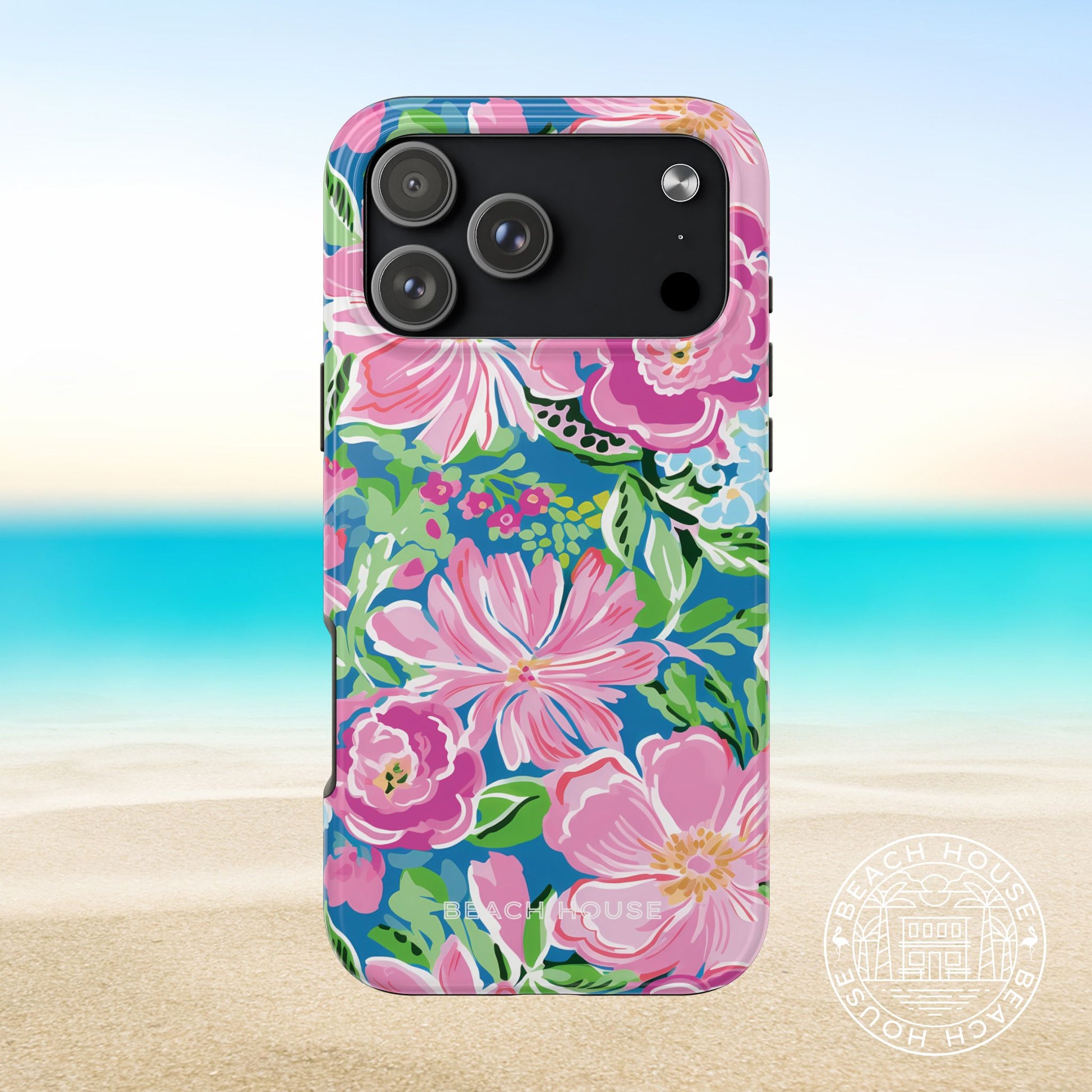 Madeira Tough Case for iPhone 17 Pro Max with pink flowers on a blue background, placed on sand with ocean view.