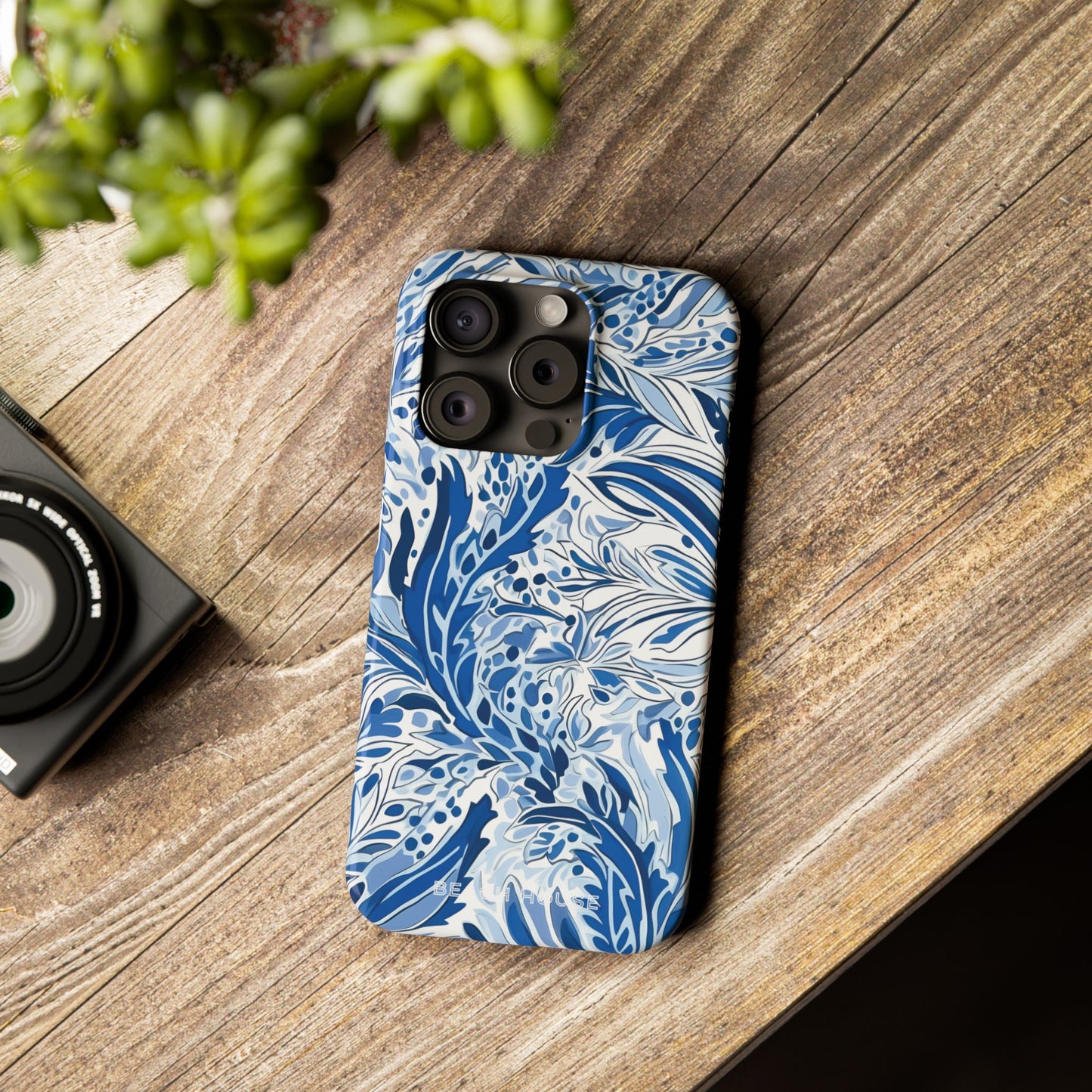 Blue floral patterned Nags Head Slim Case for iPhone on a wooden surface with a camera and plant in the background