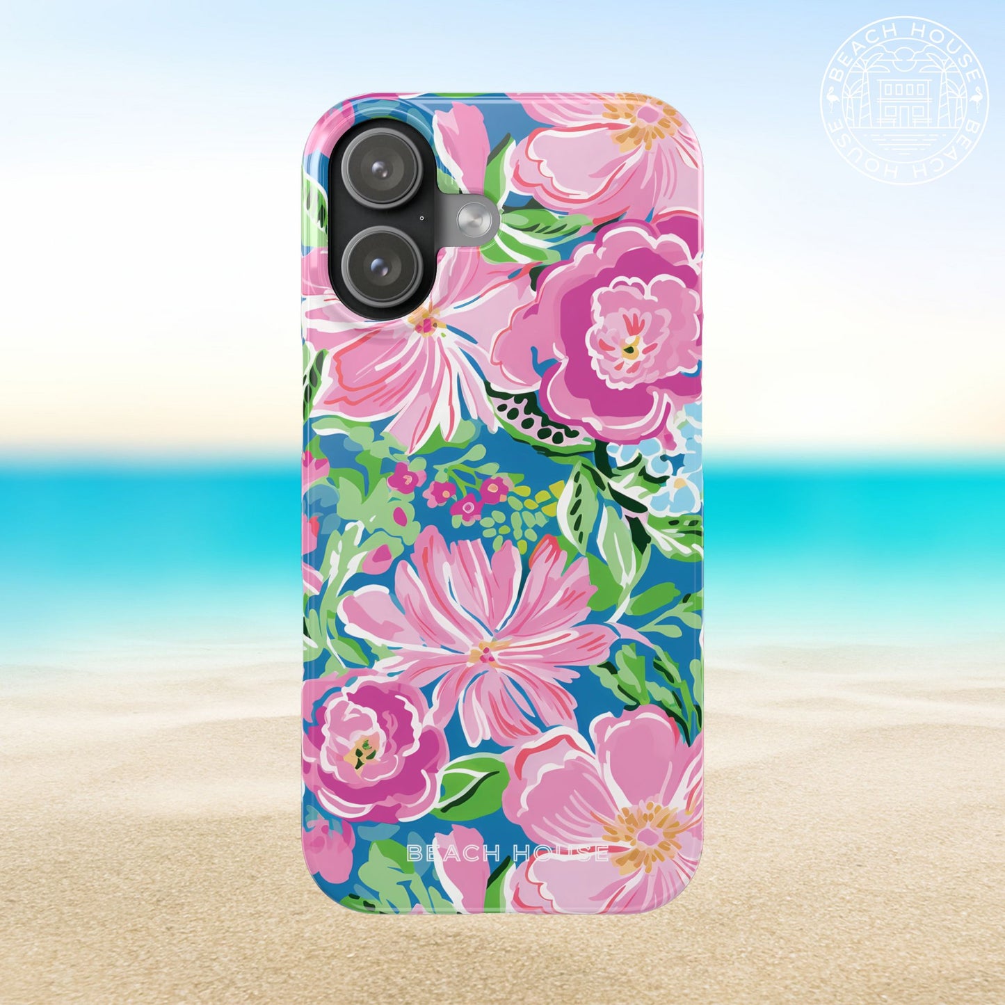 Madeira Slim Case for iPhone 17 with pink flowers on a blue background, placed on sand with ocean view.