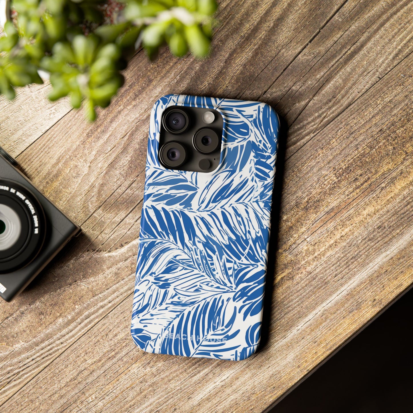 Hampton Slim Case for iPhone with blue and white leaf pattern on a wooden surface next to a camera and plant.