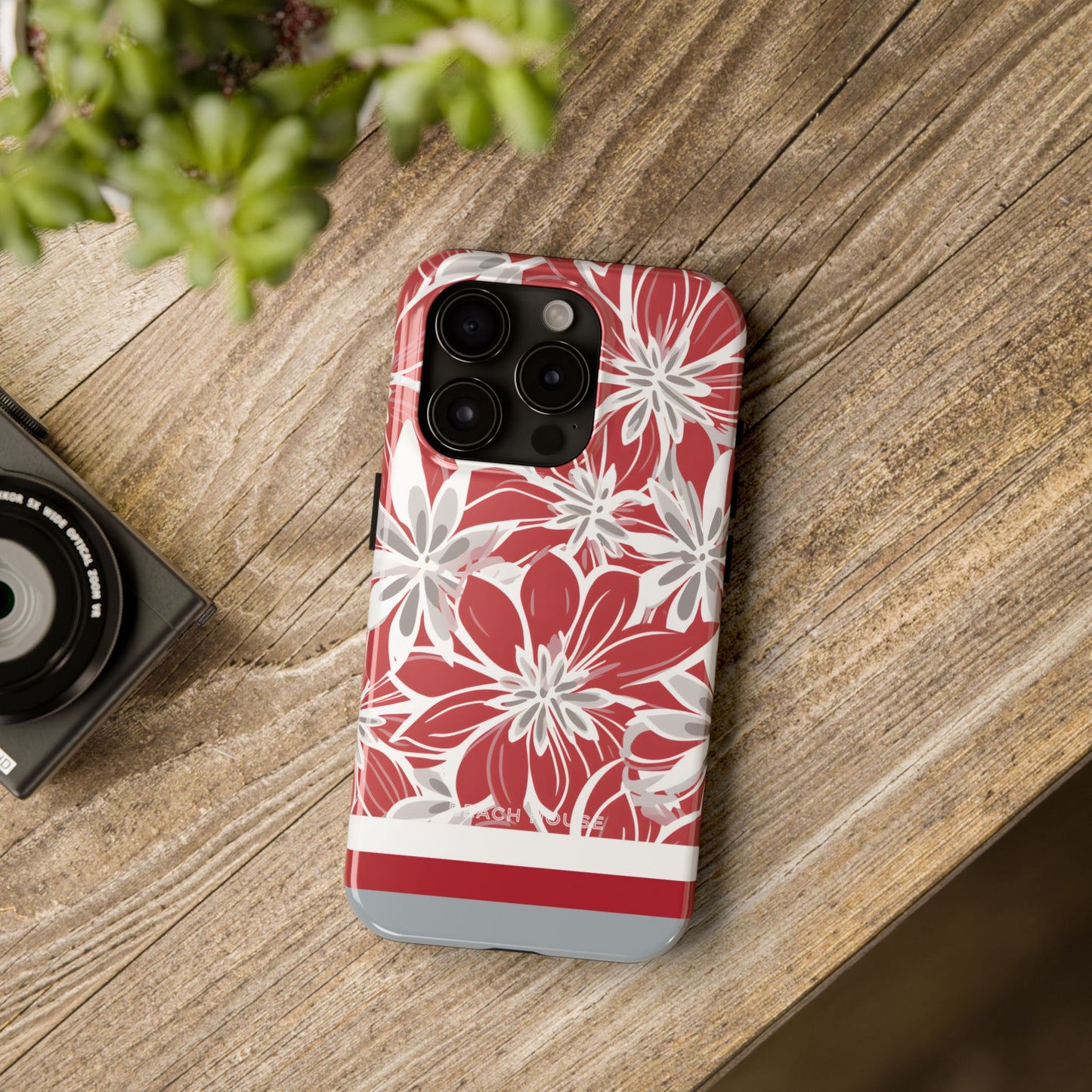 Columbus Tough case for iPhone with red and white floral pattern on a wooden surface with a camera and plant.