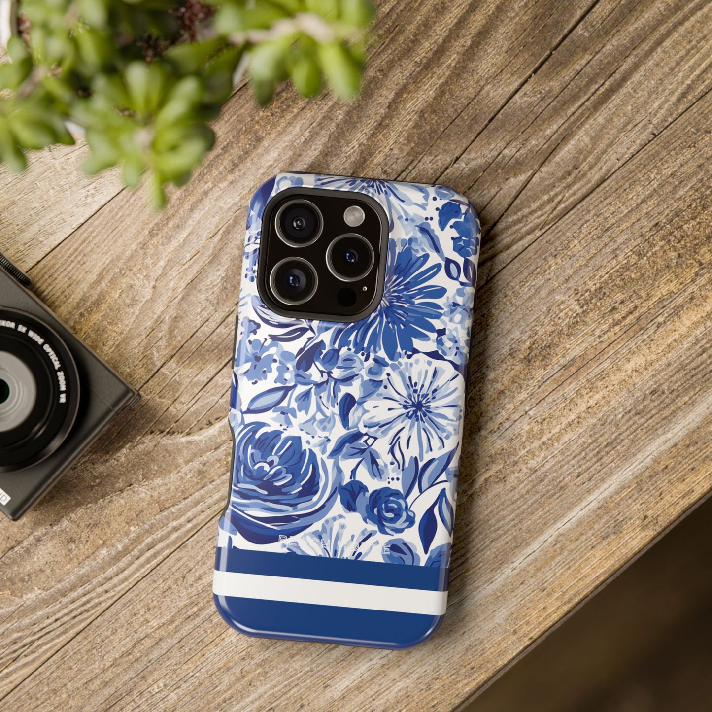 Blue floral Lexington MagSafe Tough Case for iPhone on a wooden surface with a camera and plant in the background
