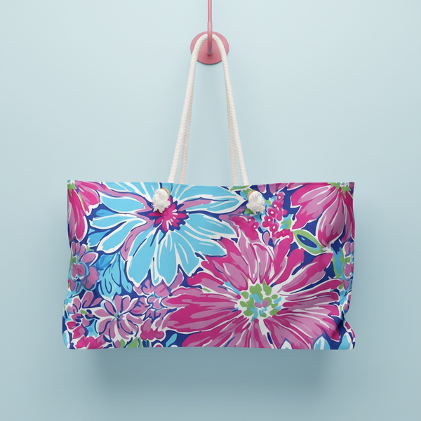 Floral patterned Bimini Weekender Tote Bag with pink and blue flowers hanging from a pink hook on a light blue background