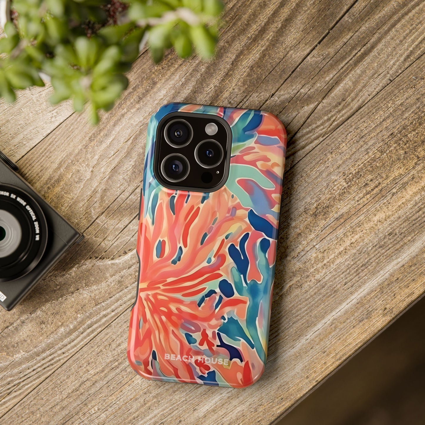 Colorful phone case with a floral pattern on a wooden surface next to a camera and plant.