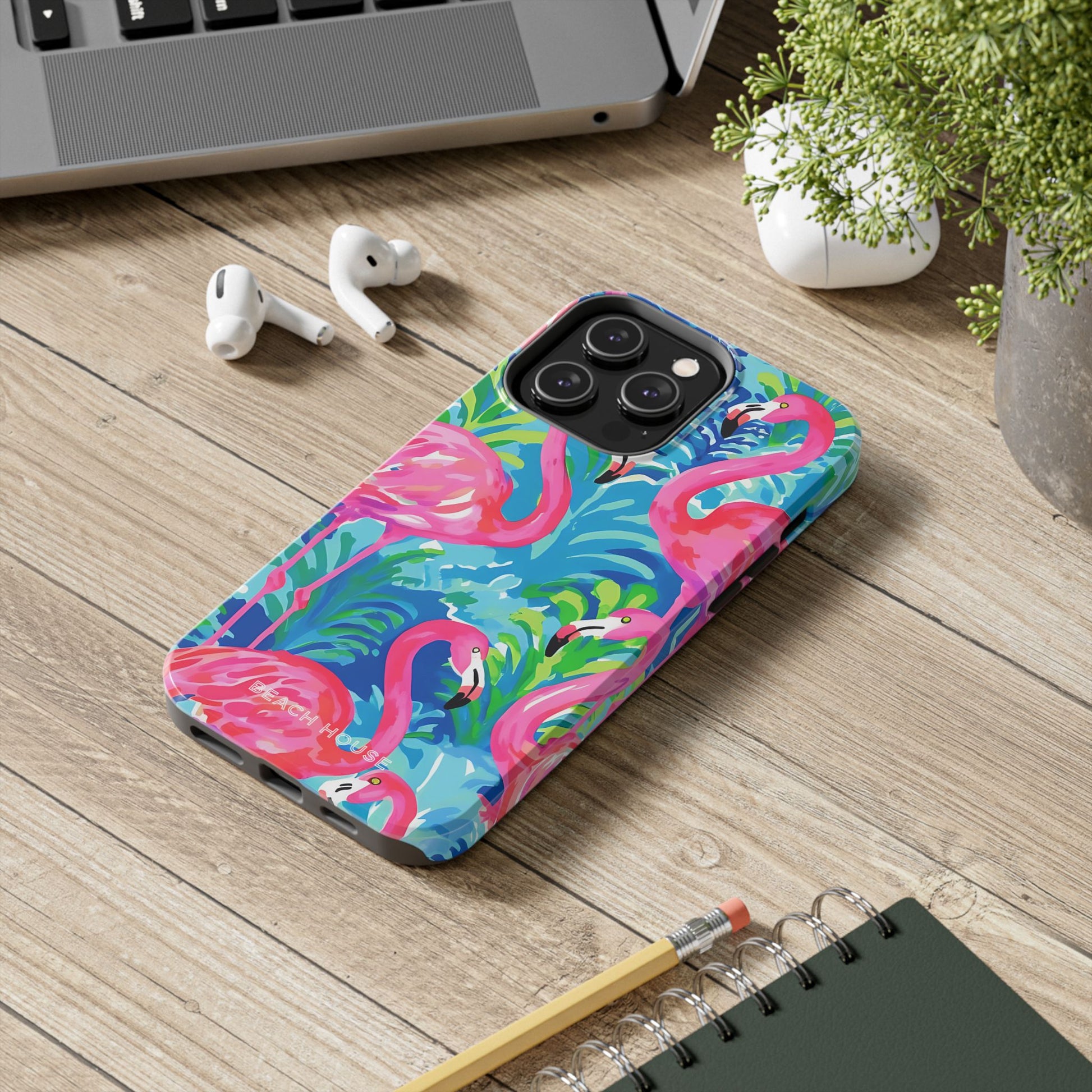 Phone case with flamingo design on a wooden surface with laptop, earbuds, and notebook.