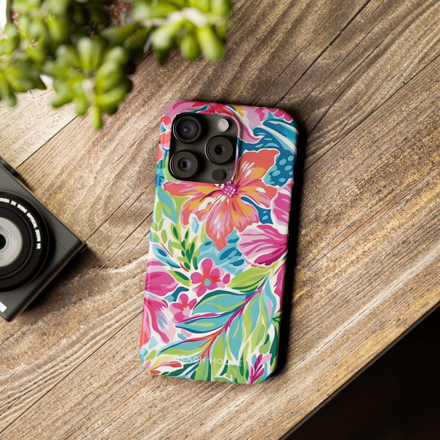Colorful Waikiki Slim Case for iPhone on a wooden surface with a camera and plant in the background