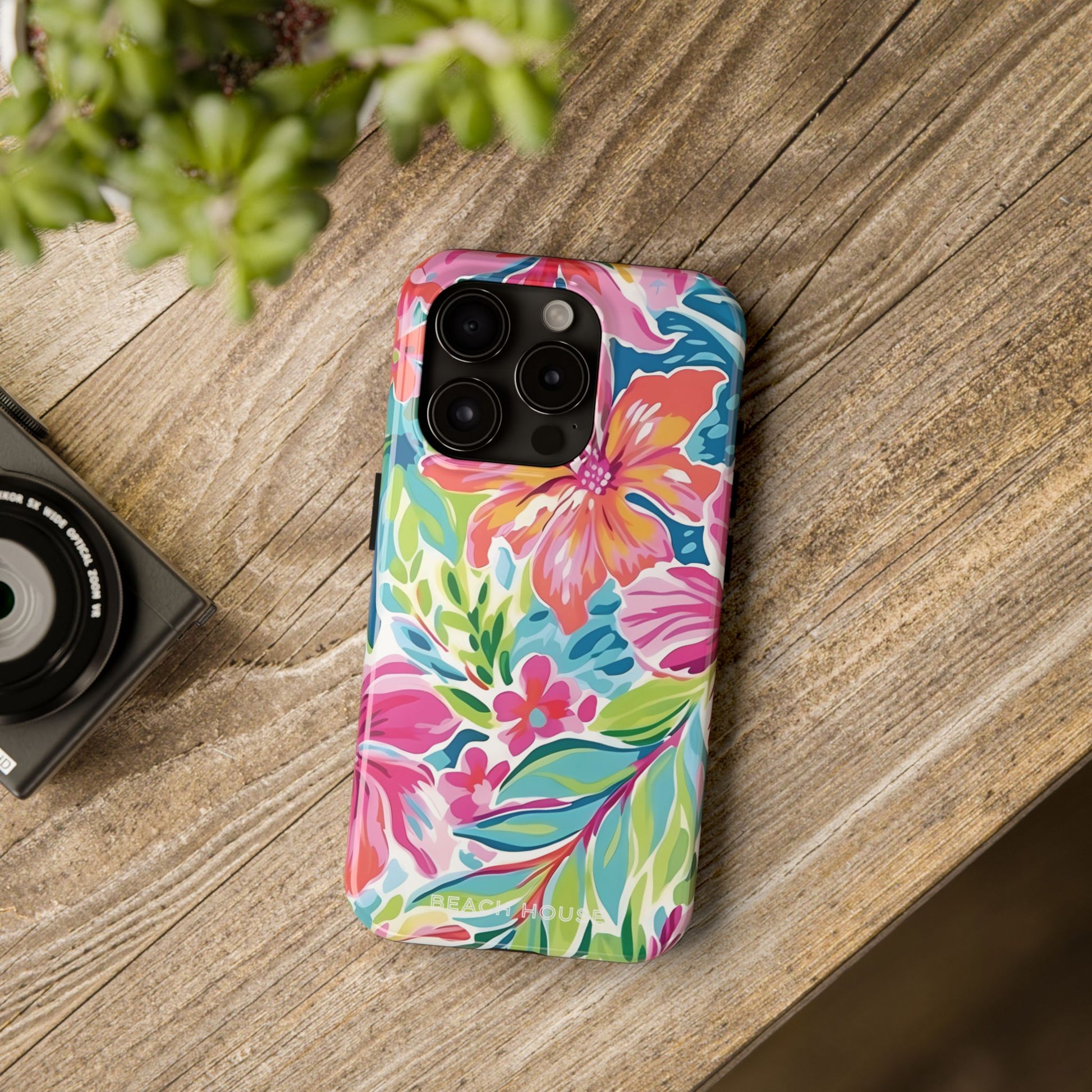 Colorful floral Waikiki Tough Case for iPhone on a wooden surface with a camera and plant in the background